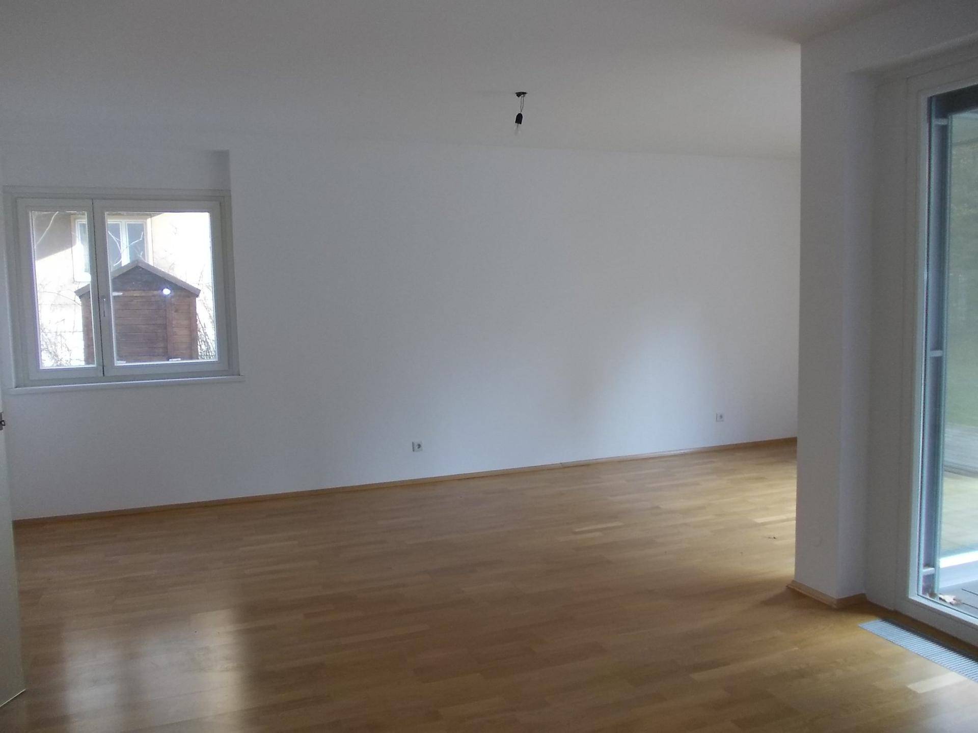 Geräumiger Wohnraum mit hellem Parkettboden und großem Fenster, das viel Tageslicht hereinlässt.