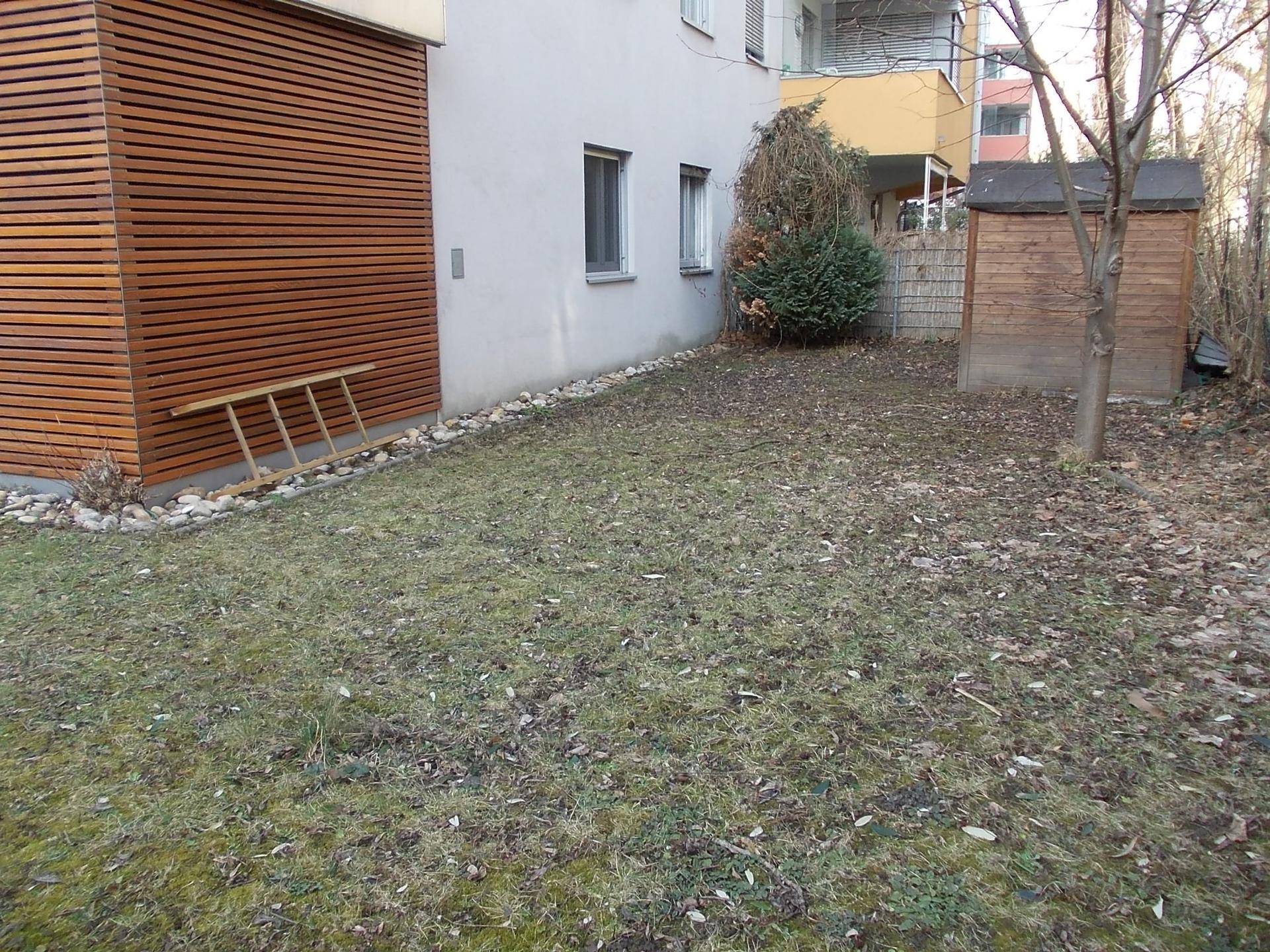 Gartenbereich mit Rasen, einem kleinen Holzschuppen und der Seitenansicht des Gebäudes.