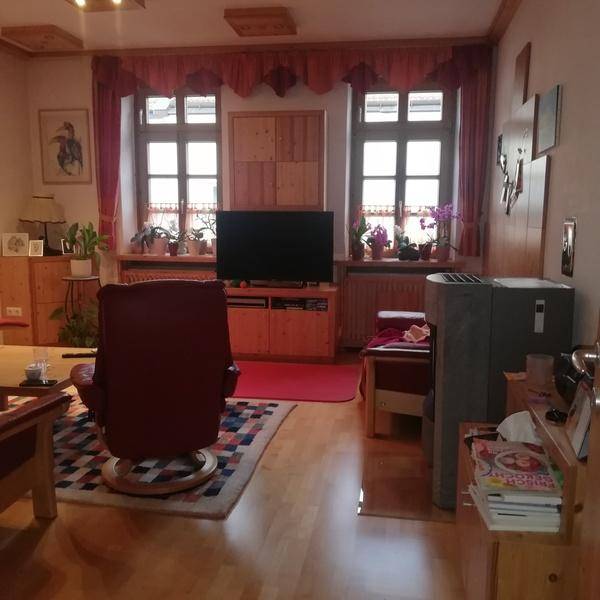 Geräumiges Wohnzimmer mit Holzboden, zwei Fenstern und einem bequemen Sessel.