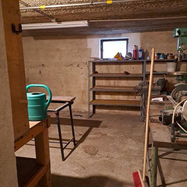 Werkstatt im Keller mit Arbeitsflächen, Regalen und Werkzeugen, gut beleuchtet durch ein kleines Fenster.