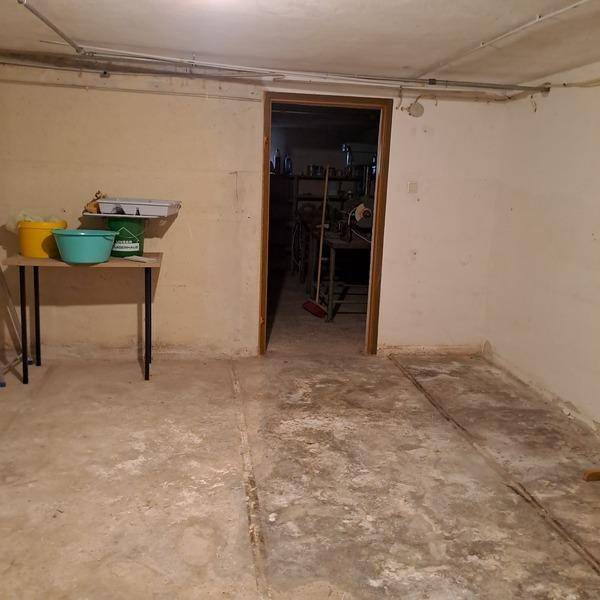 Geräumiger Kellerflur mit Betonboden und Zugang zu weiteren Kellerräumen, mit sichtbaren Leitungen.