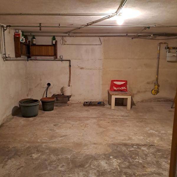 Unausgebauter Kellerraum mit Betonboden, sichtbaren Rohren und einfacher Beleuchtung.