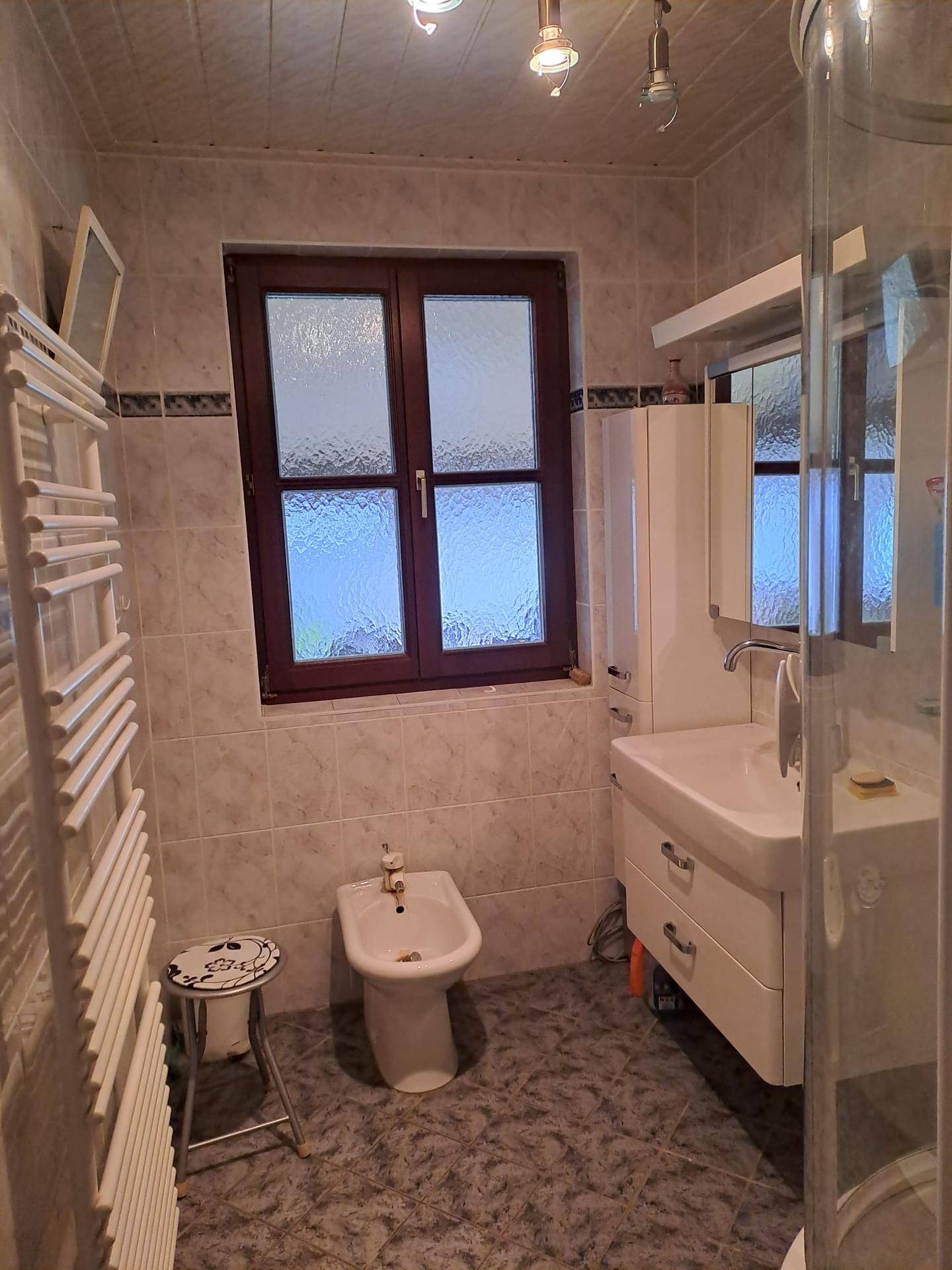 Gefliestes Badezimmer mit Fenster, Bidet, Waschbecken und Duschkabine.