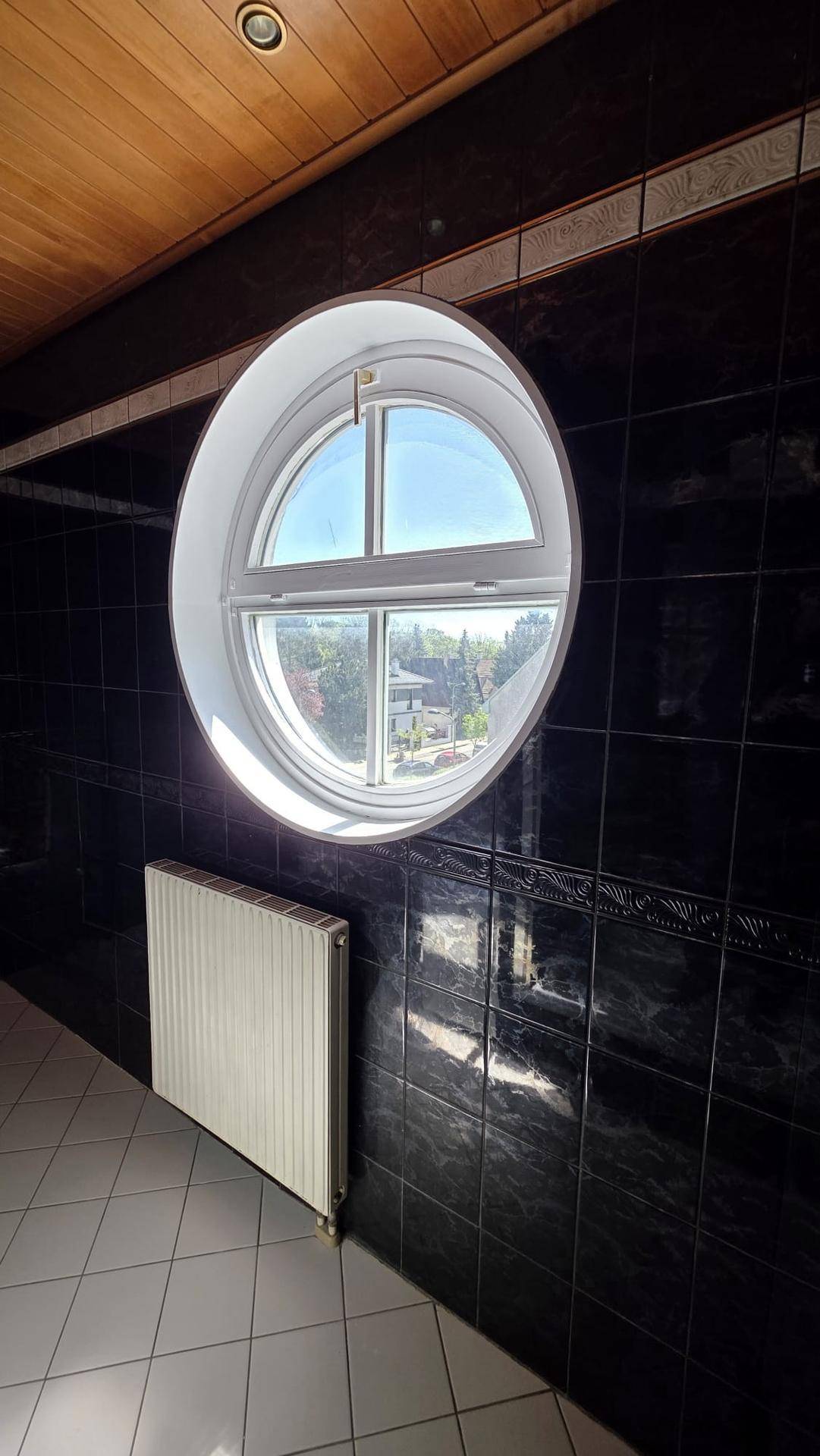 Badezimmer mit dunklen Fliesen, rundem Fenster, Heizkörper und Holzdecke.