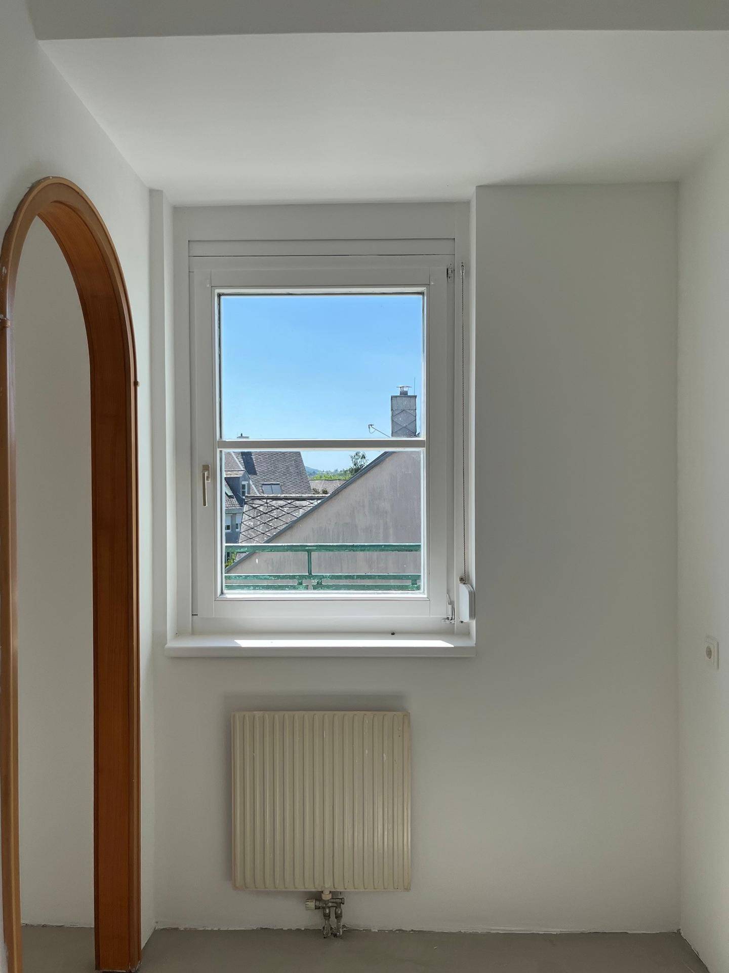 Leerer Raum mit Fenster, Heizkörper und Blick auf die umliegenden Dächer.