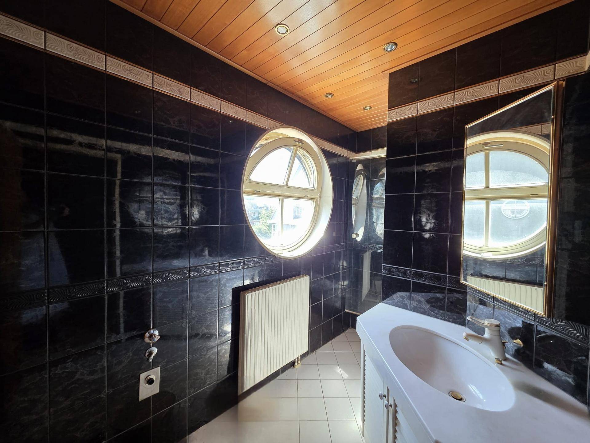 Badezimmer mit dunklen Fliesen, rundem Fenster, Waschtisch und Holzdecke.