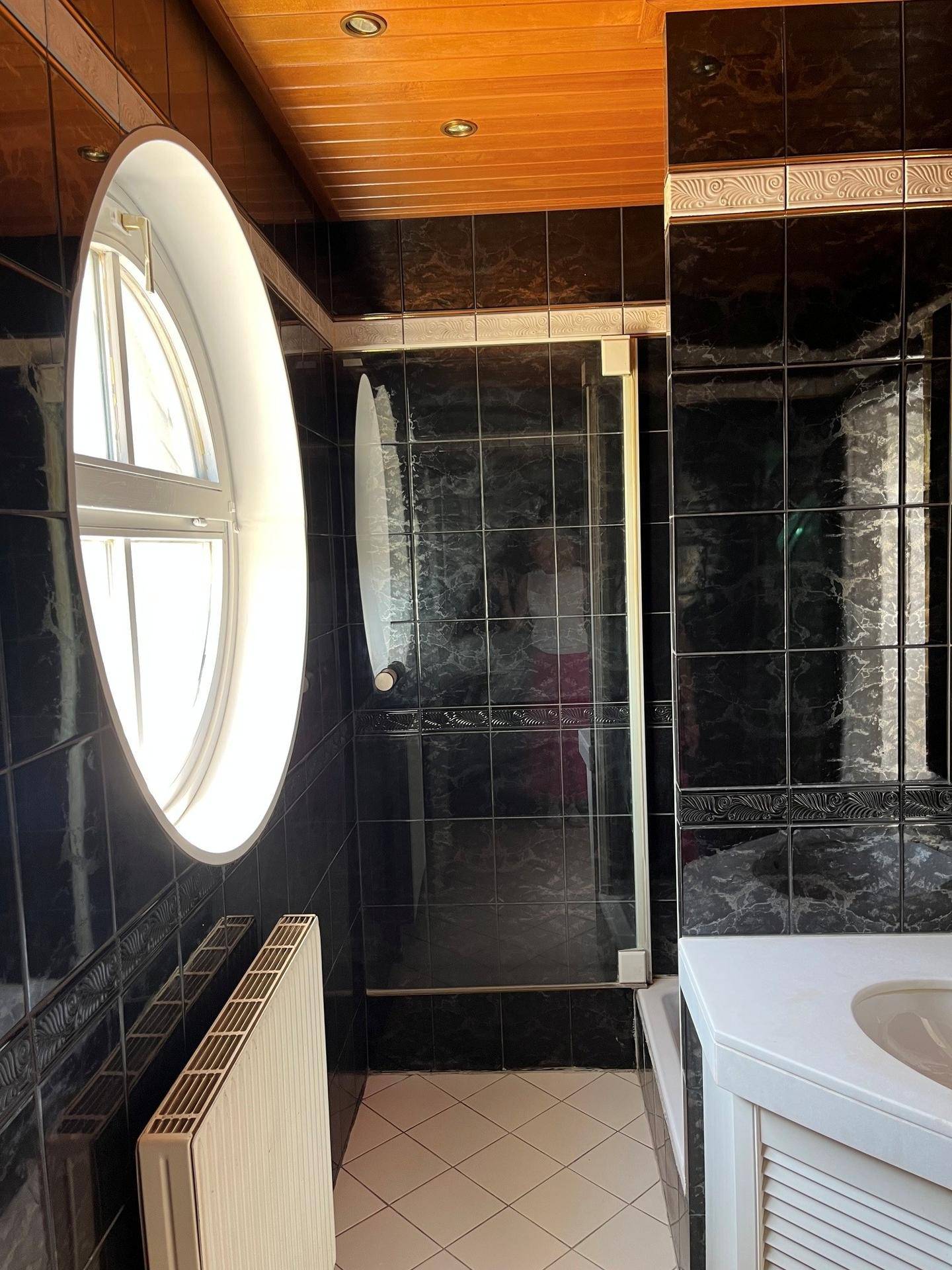 Badezimmer mit dunklen Fliesen, rundem Fenster, Duschbereich und Holzdecke.