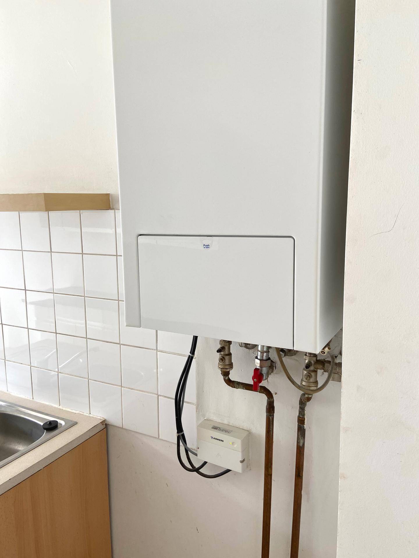 Nahaufnahme eines Gasboilers an einer gefliesten Wand, mit sichtbaren Rohren und Anschlüssen.