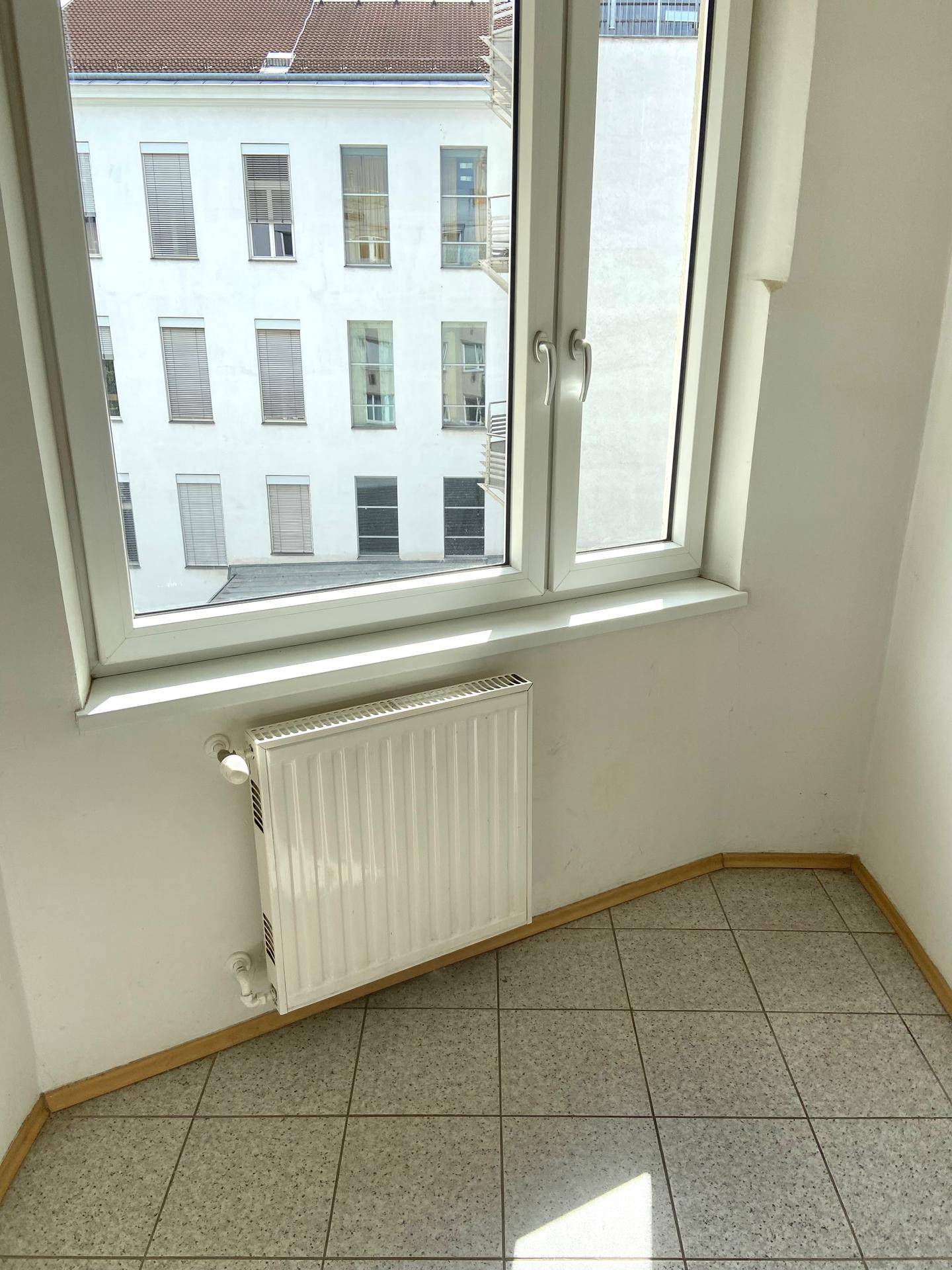 Heller Raumeck mit Fenster und Heizkörper, gefliestem Boden und weiß gestrichenen Wänden.
