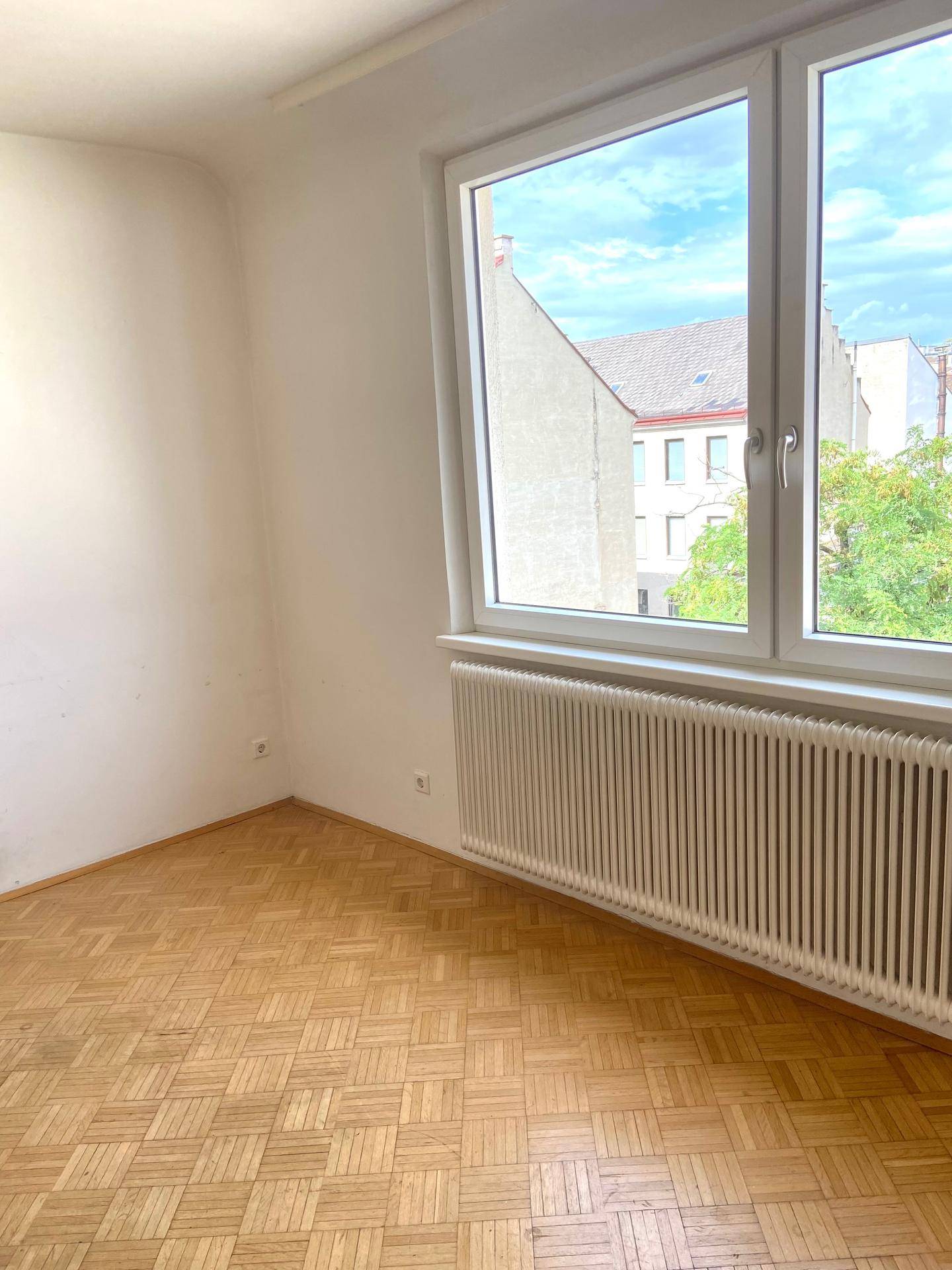 Heller, leerer Raum mit Fischgrätparkettboden, großem Fenster und Heizkörper.