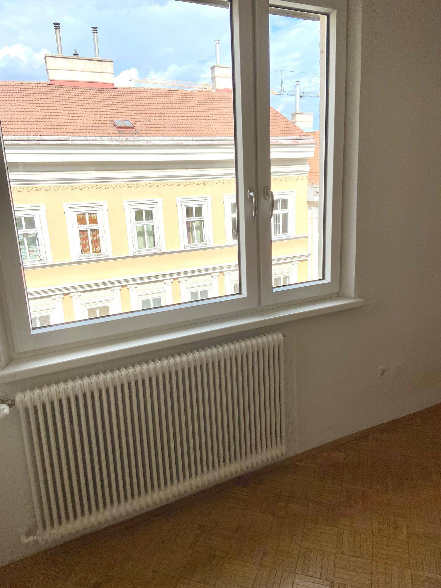 Heller Raum mit Fischgrätparkettboden, Fenster und Heizkörper, Blick auf ein Altbaugebäude.