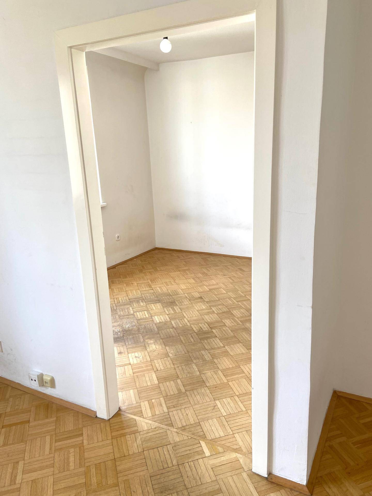 Blick durch eine Tür in einen leeren Raum mit Fischgrätparkettboden und weißen Wänden.