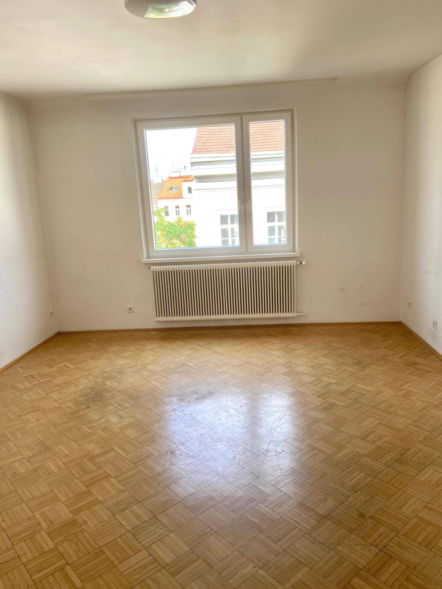 Geräumiger, leerer Raum mit Fischgrätparkettboden, großem Fenster und Heizkörper.