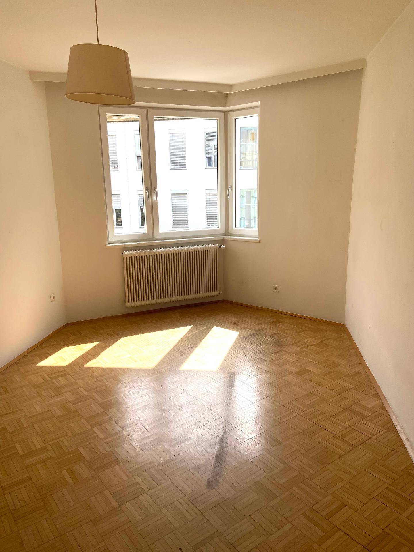 Heller, leerer Raum mit Fischgrätparkettboden, Erkerfenster und viel natürlichem Licht.