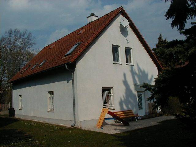 Seitenansicht eines weißen Hauses mit rotem Ziegeldach, Dachfenstern und Satellitenschüssel im Grünen.