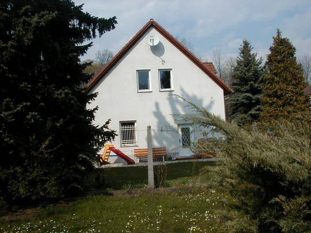 Vorderansicht des weißen Hauses mit rotem Dach, umgeben von hohen Bäumen und einem kleinen Gartenbereich.