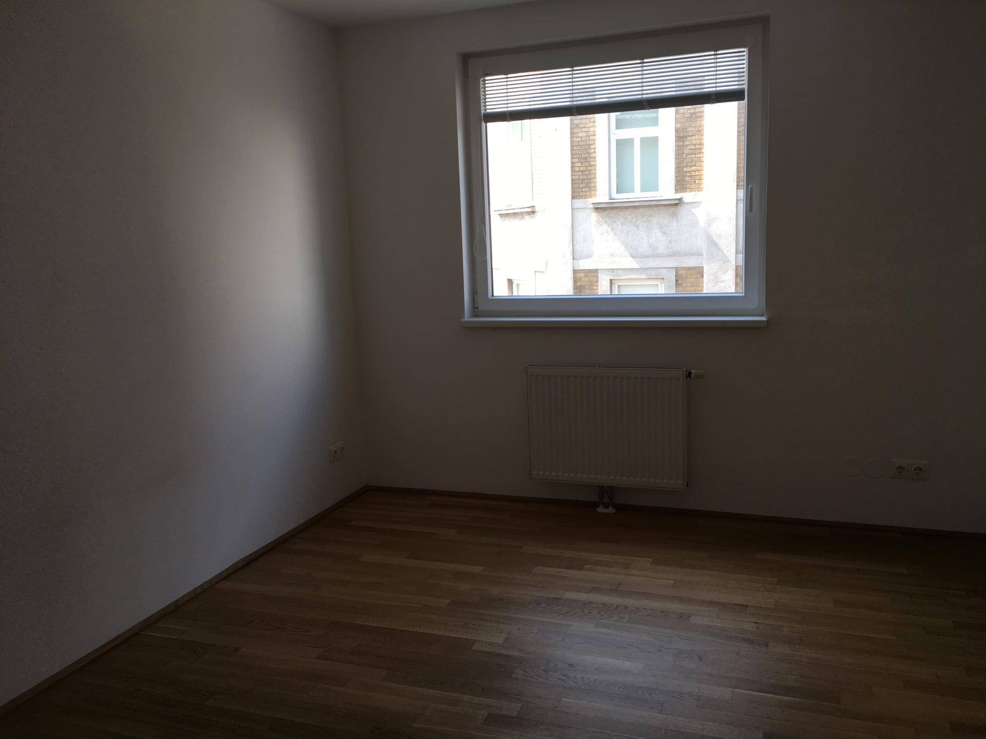 Leeres Zimmer mit Parkettboden, einem Fenster und Heizkörper, bietet Blick auf ein gegenüberliegendes Gebäude.