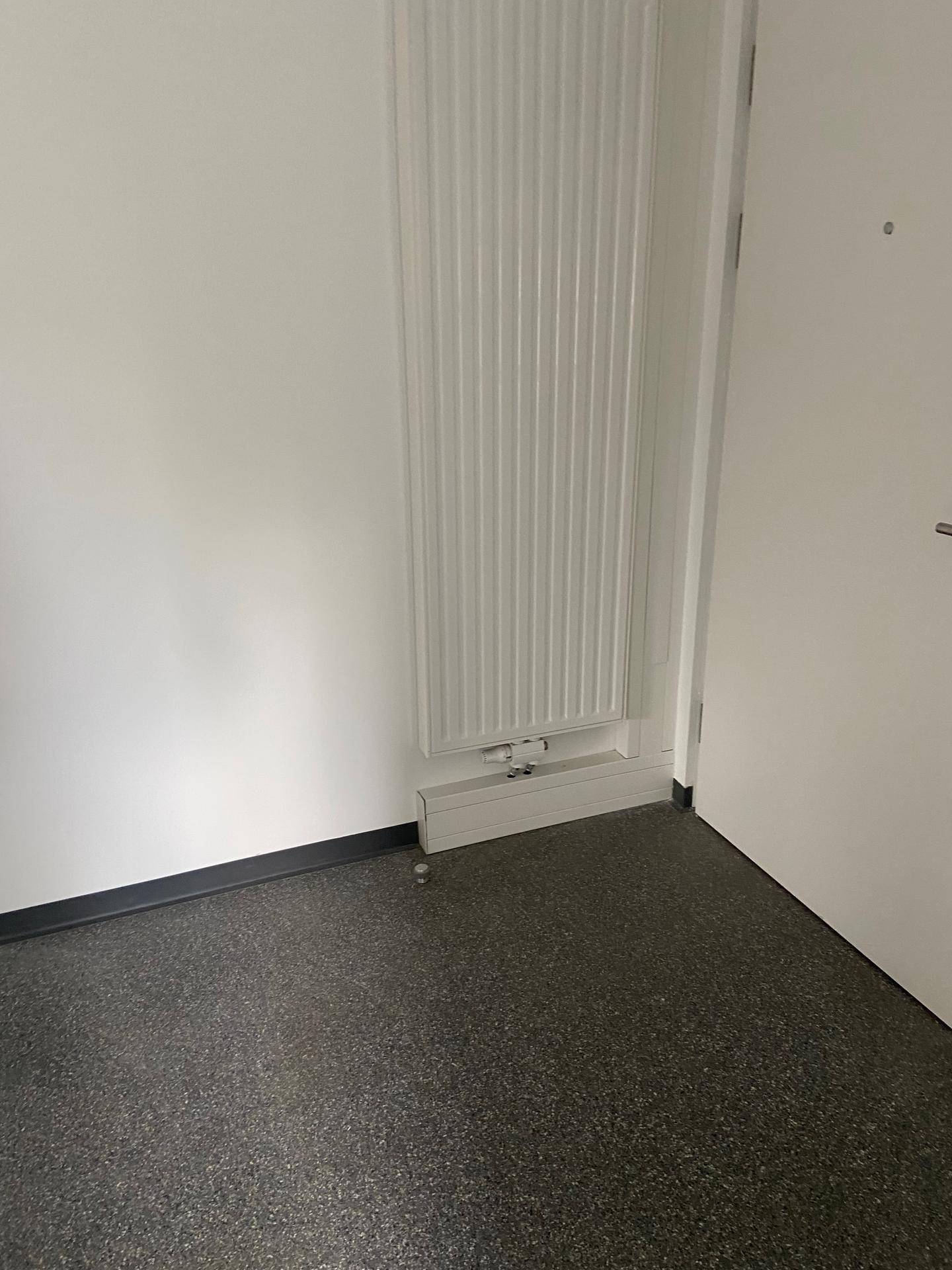 Detailansicht eines weißen Heizkörpers an einer weißen Wand über grauem Vinylboden.