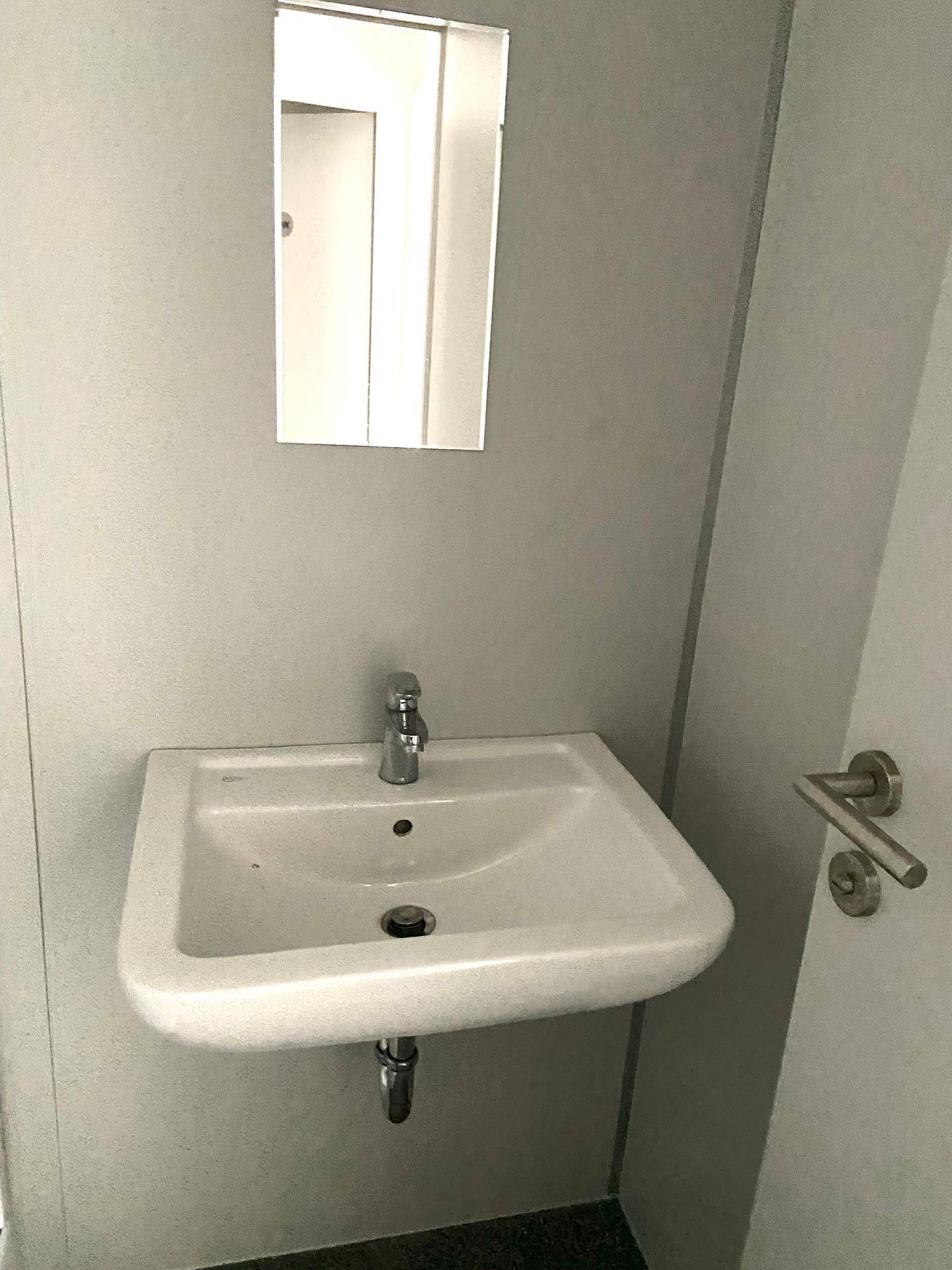 Kleines Waschbecken mit Spiegel an einer hellgrauen Wand in einem Badezimmer.