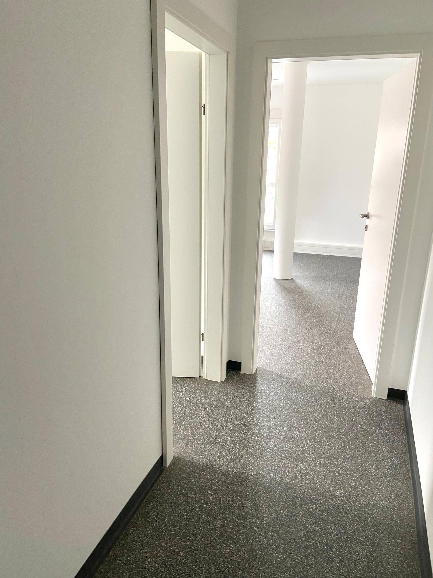 Heller Flur mit grauem Vinylboden und zwei offenen Türen, die zu weiteren Räumen führen.