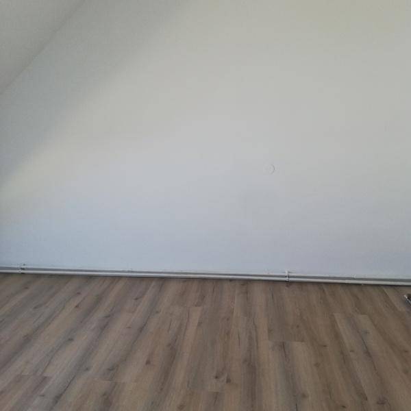 Leerer Raum mit weißer Wand, Laminatboden und sichtbaren Heizungsrohren.