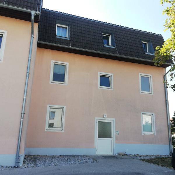 Mehrfamilienhaus mit hellrosa Fassade, dunklem Mansarddach und mehreren Fenstern, umgeben von Grün.