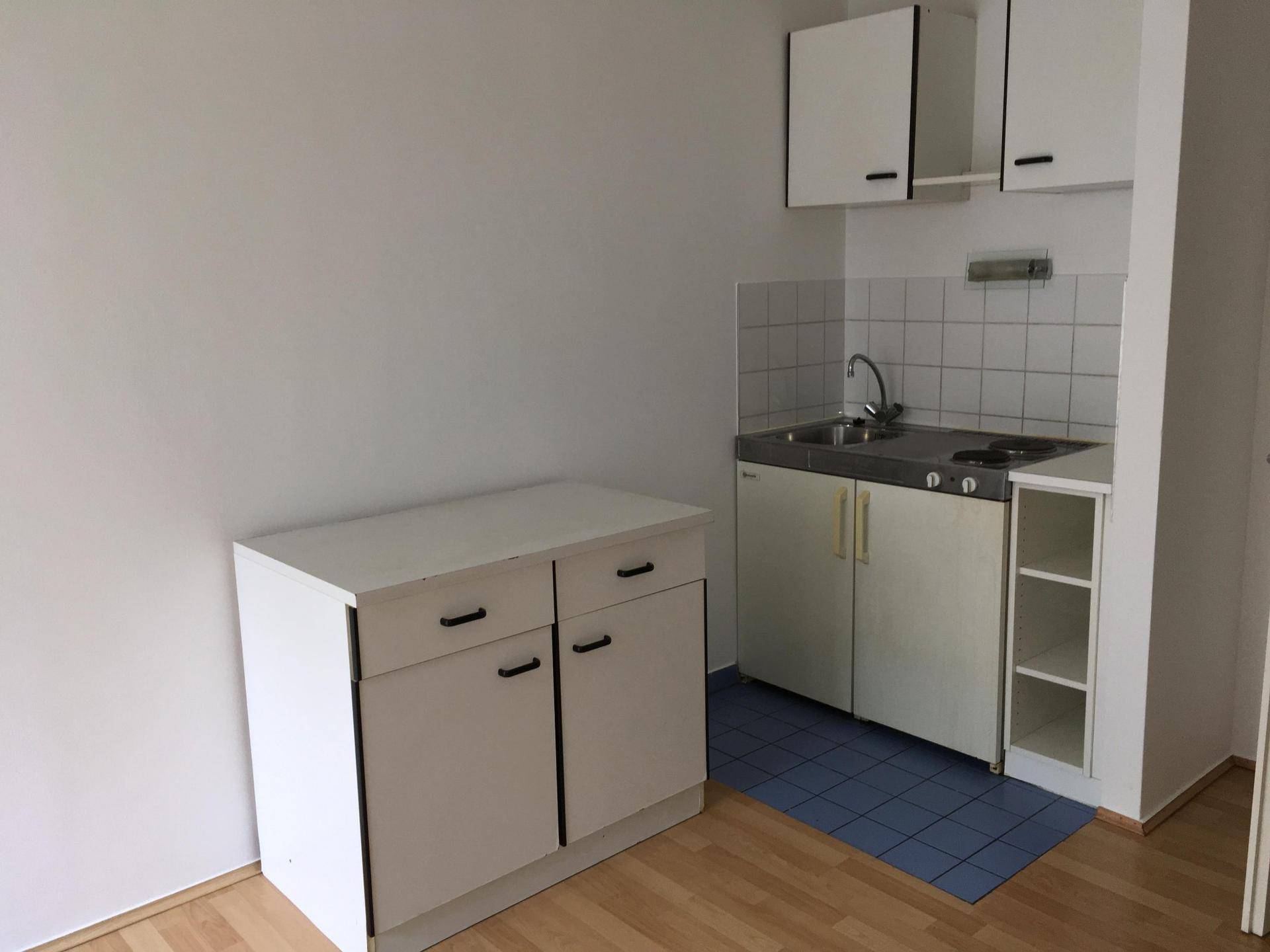 Kompakte Küchenzeile mit weißen Schränken, Spüle, Kochfeld und Kühlschrank auf blau gefliestem Boden.