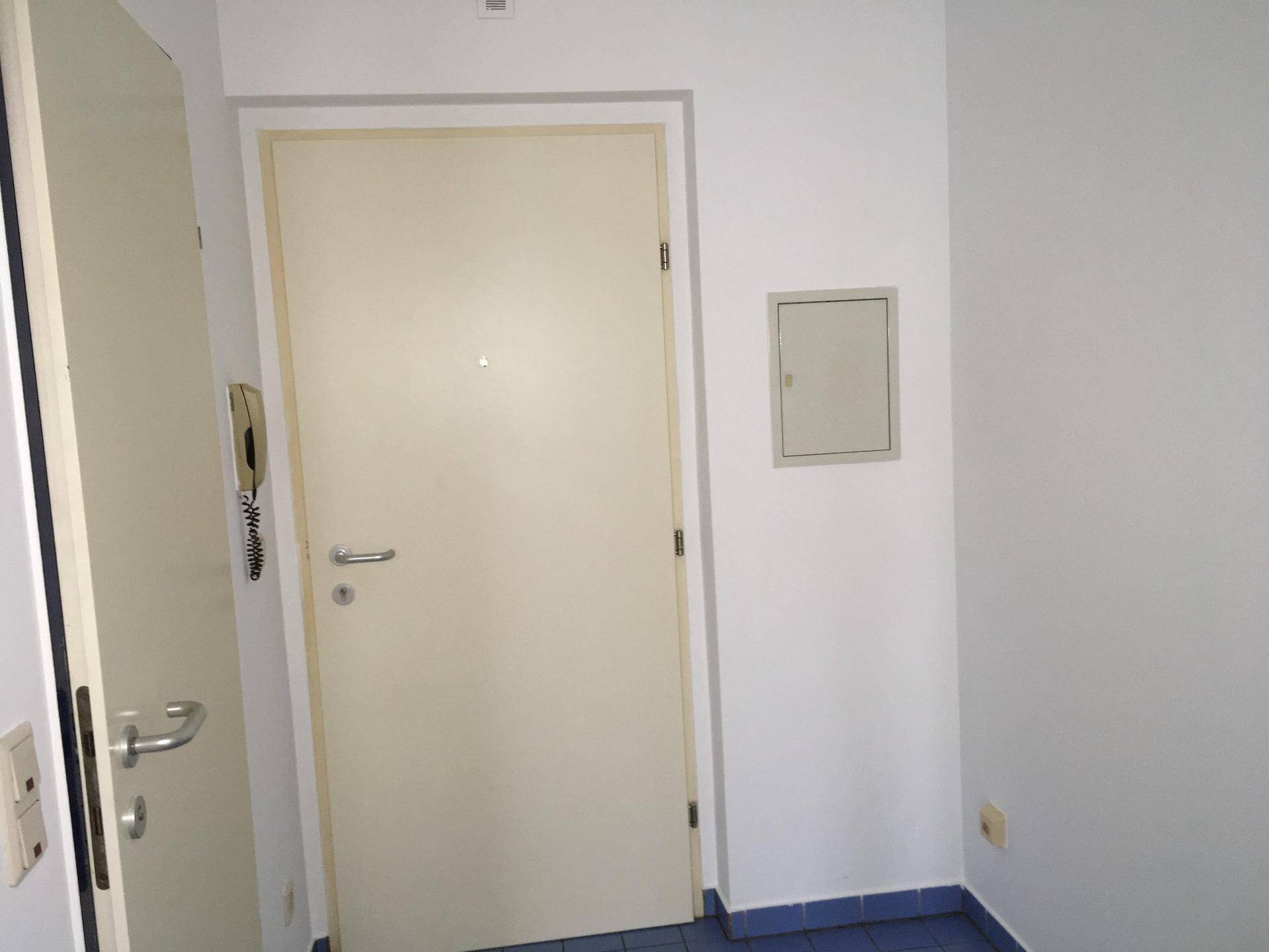 Eingangsbereich mit heller Tür, Gegensprechanlage und blau gefliestem Boden, der einen praktischen Empfang bietet.