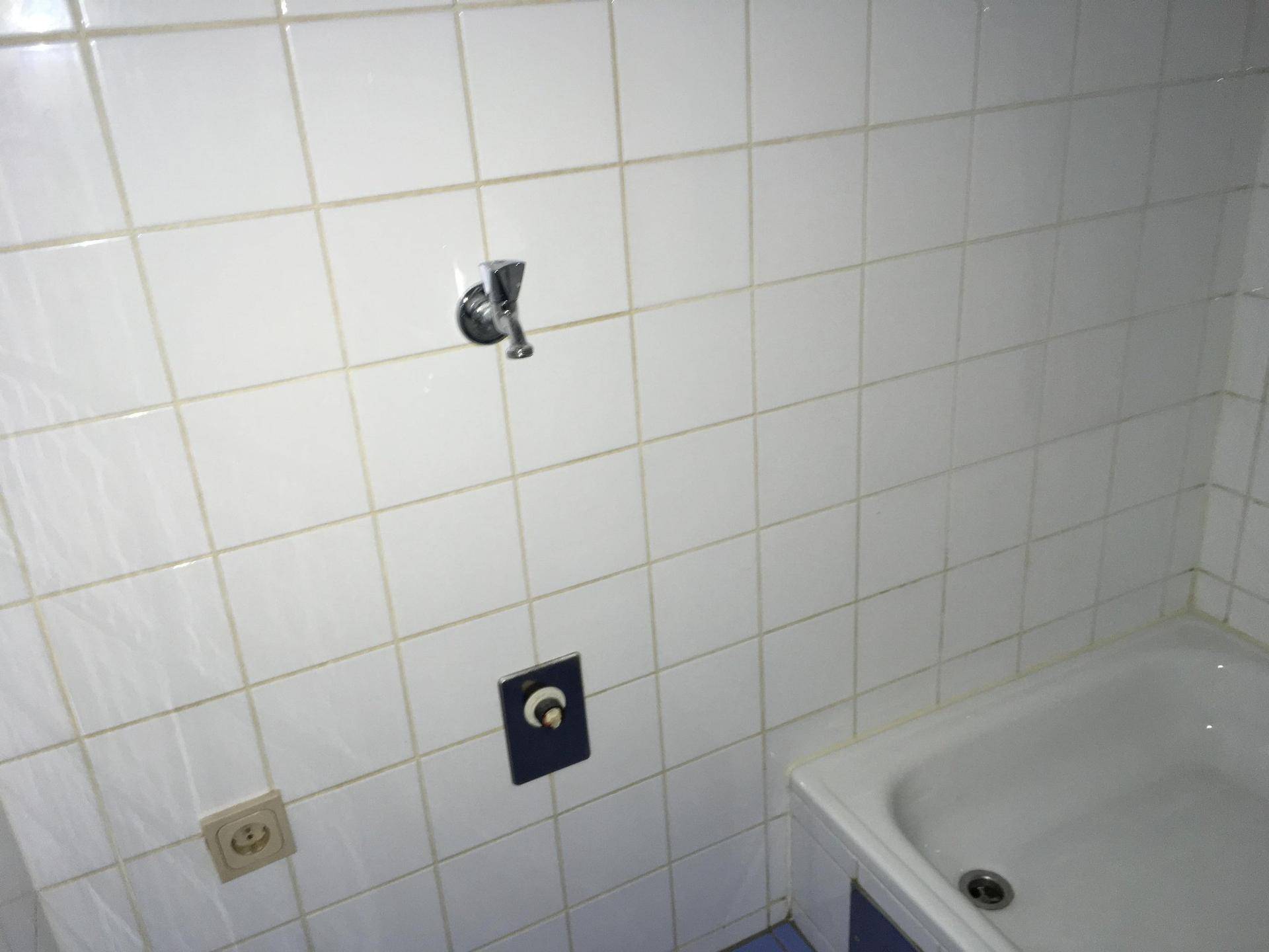 Badezimmerwand mit weißen Fliesen und Armaturen für eine Badewanne, die auf eine einfache Ausstattung hinweist.