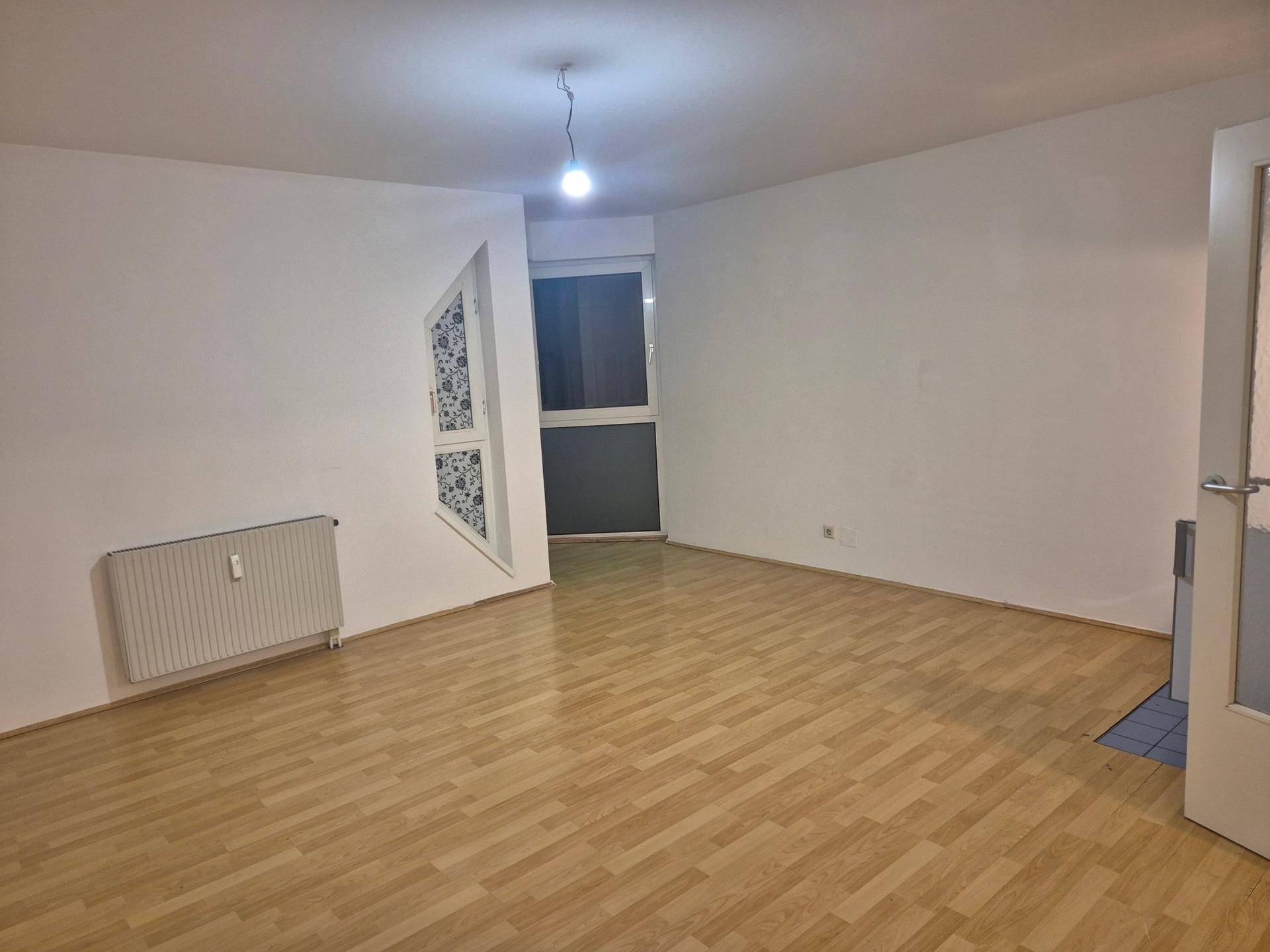 Geräumiger, leerer Raum mit Parkettboden, Heizkörper und einem Fenster mit gemustertem Vorhang.