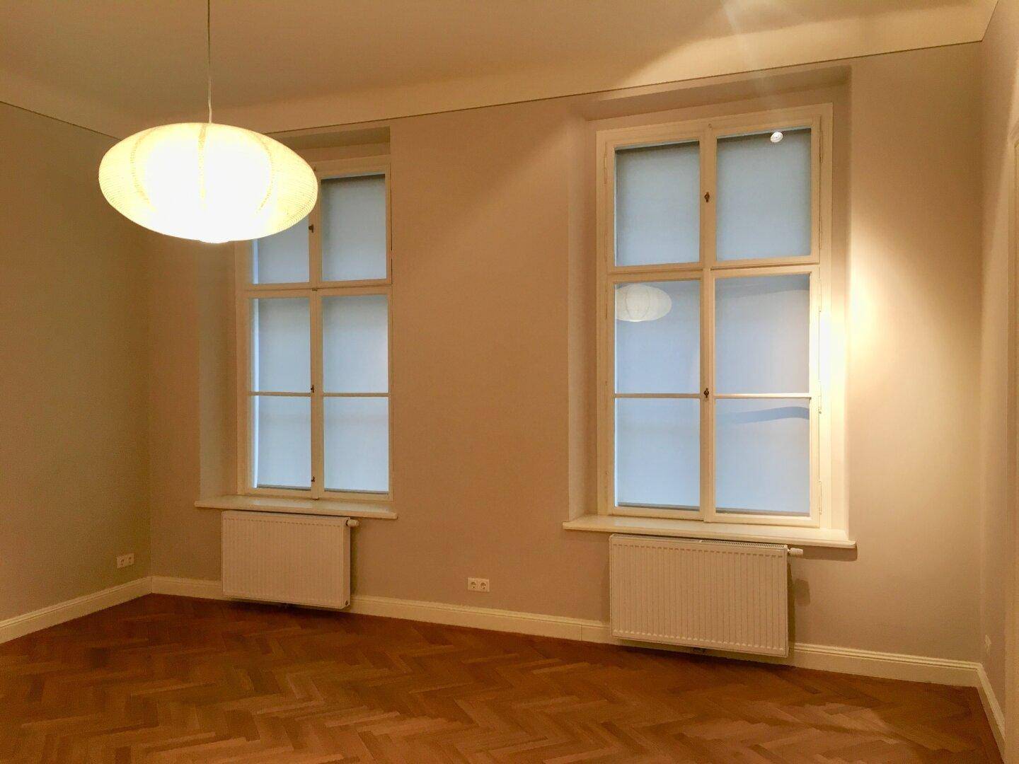 Helles Zimmer mit zwei hohen Fenstern, Heizkörpern und schönem Fischgrätparkettboden.