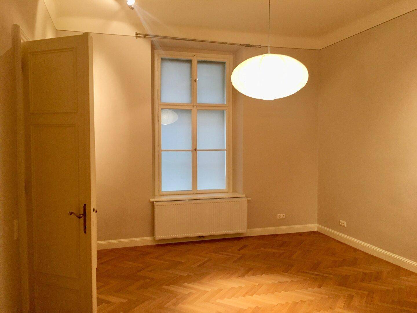 Heller Raum mit einem hohen Fenster, Heizkörper und elegantem Fischgrätparkettboden.