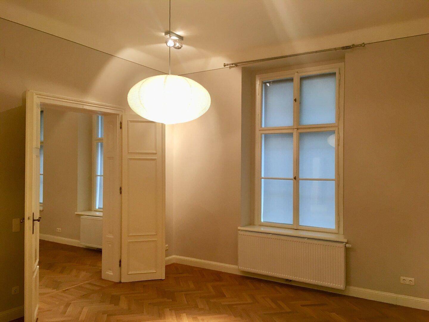 Geräumiges Zimmer mit Fischgrätparkett, einem hohen Fenster und einem Durchgang zu einem weiteren Raum.