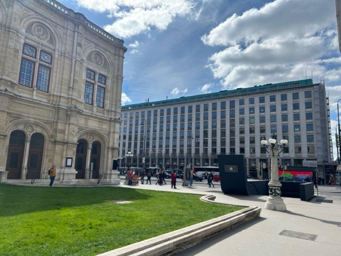 Historisches Gebäude mit aufwendiger Fassade neben einem modernen Bau an einer belebten Straße mit Grünfläche.