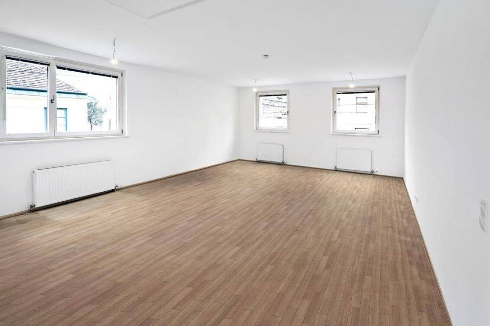 Großer, leerer Raum mit Holzboden und mehreren Fenstern, die viel Tageslicht hereinlassen und eine ruhige Umgebung zeigen.