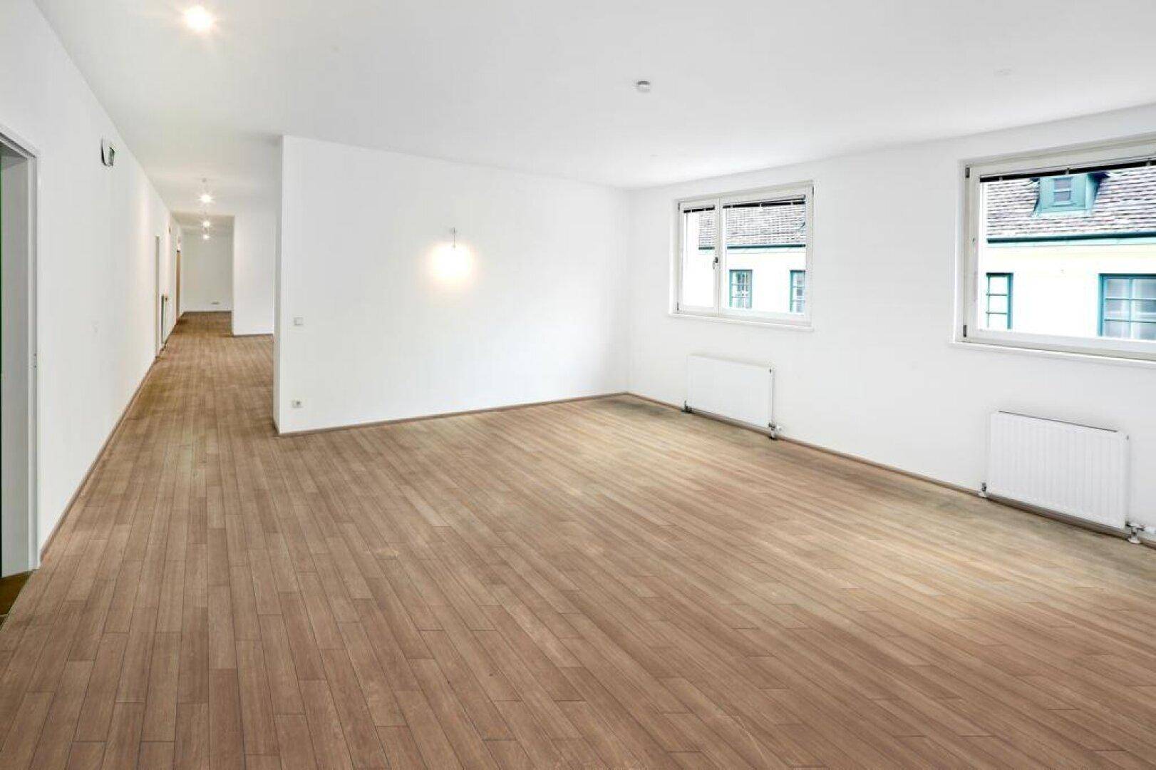 Geräumiger, heller Raum mit Holzboden und großen Fenstern, die einen Blick auf die umliegenden Gebäude bieten.