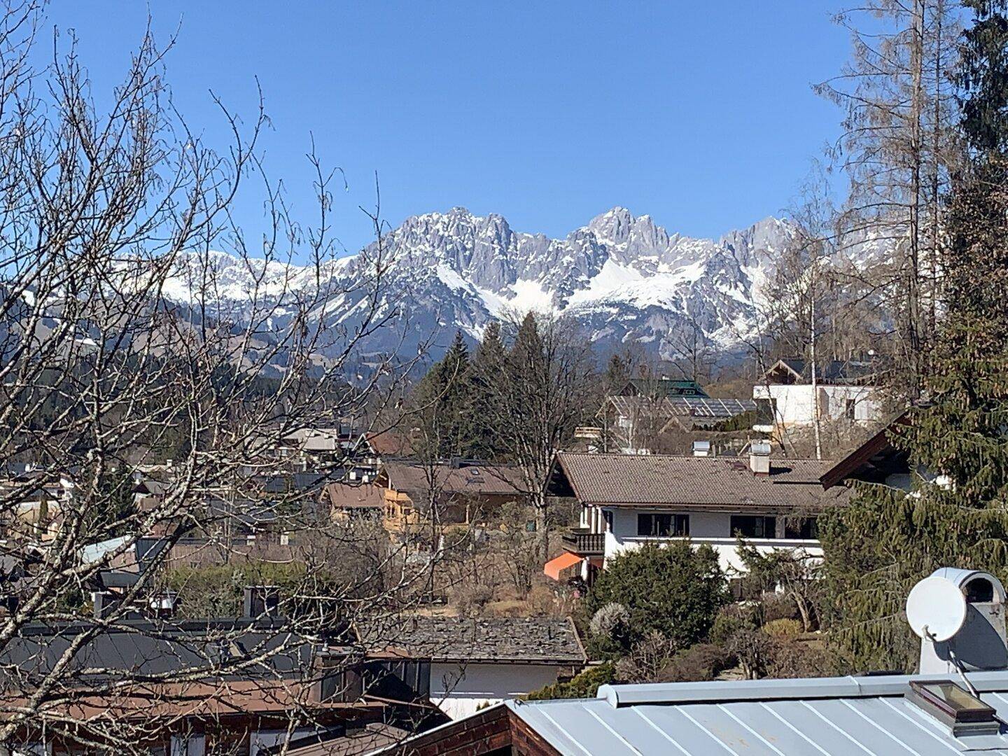 Sonniger Blick auf eine verschneite Bergkette über den Dächern einer Stadt mit kahlen Bäumen im Vordergrund.