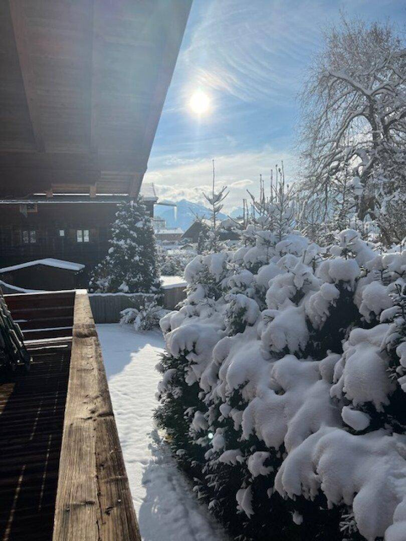 Sonniger Wintertag mit schneebedeckten Büschen und einem Holzbalkon, im Hintergrund sind Berge sichtbar.