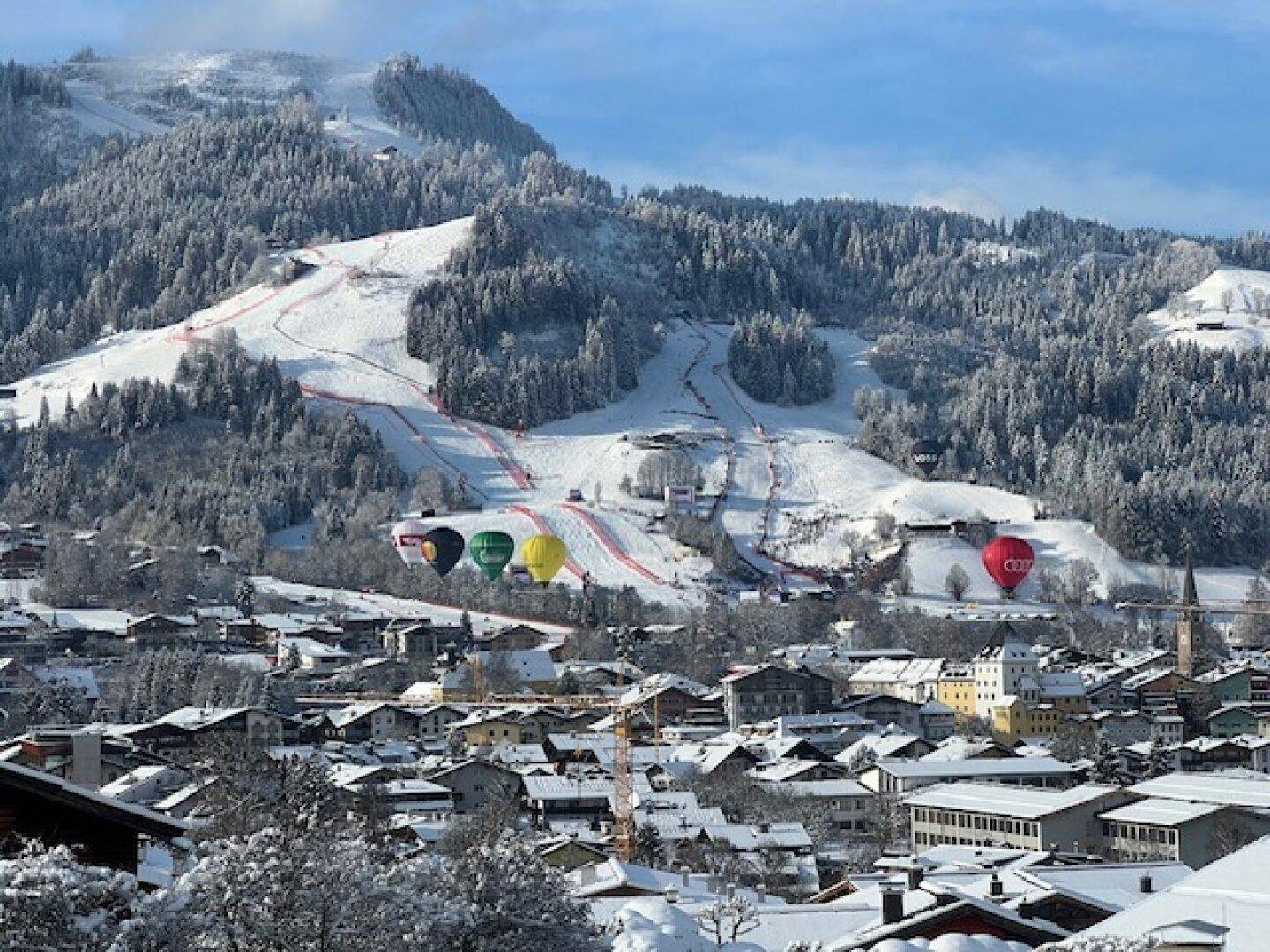Panoramablick auf eine verschneite Stadt mit Skipisten und Bergen im Hintergrund, Heißluftballons am Himmel.