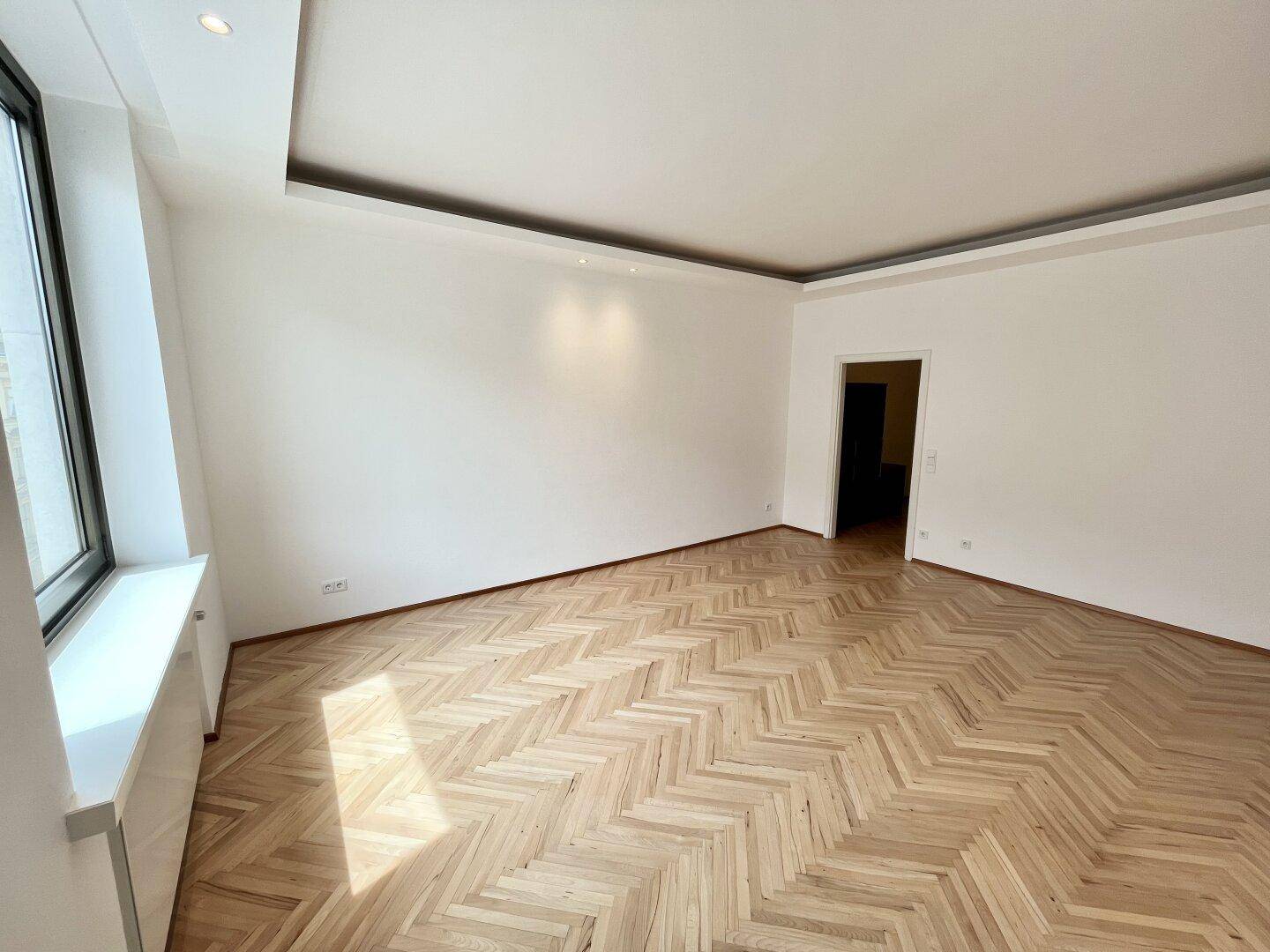 Heller Raum mit modernem Fischgrätparkett, großem Fenster und indirekter Deckenbeleuchtung.