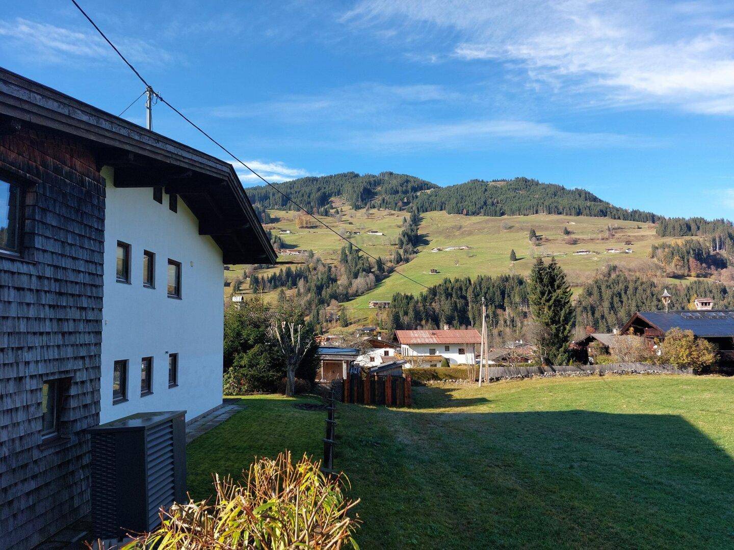 Seitenansicht eines Hauses mit Blick auf weite grüne Wiesen und bewaldete Berghänge unter blauem Himmel.