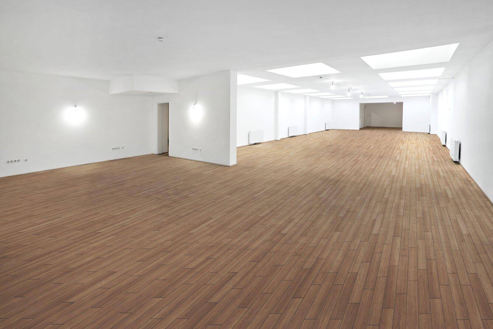 Großer, leerer Raum mit Holzboden, weißen Wänden und mehreren Deckenfenstern, die für Helligkeit sorgen.