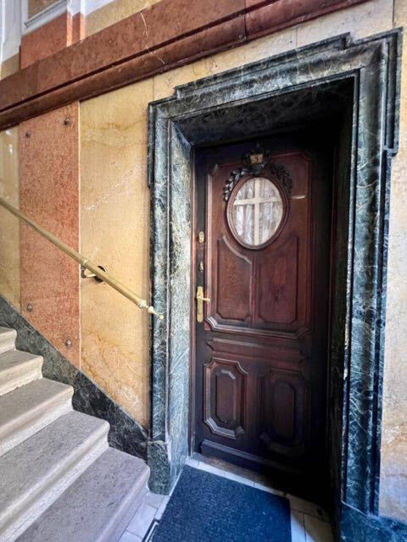Antike Holztür mit rundem Fenster, umrahmt von dunklem Marmor, neben einer Steintreppe.