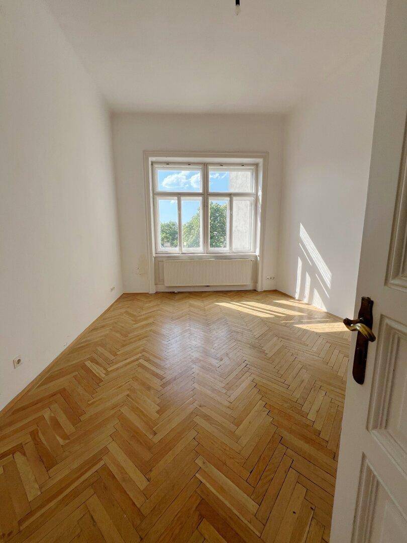 Helles Zimmer mit klassischem Fischgrätparkett und einem großen Fenster mit viel Tageslicht.