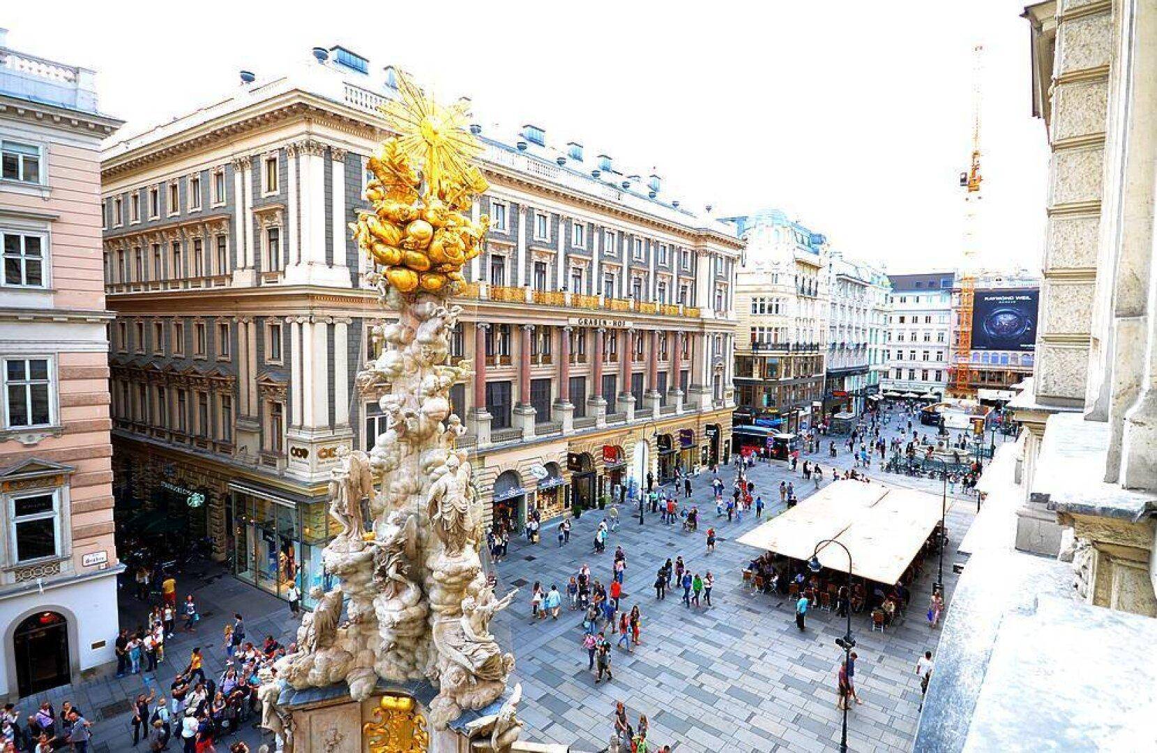 Blick auf eine belebte Stadtstraße mit historischen Gebäuden und einem Denkmal, umgeben von Geschäften.