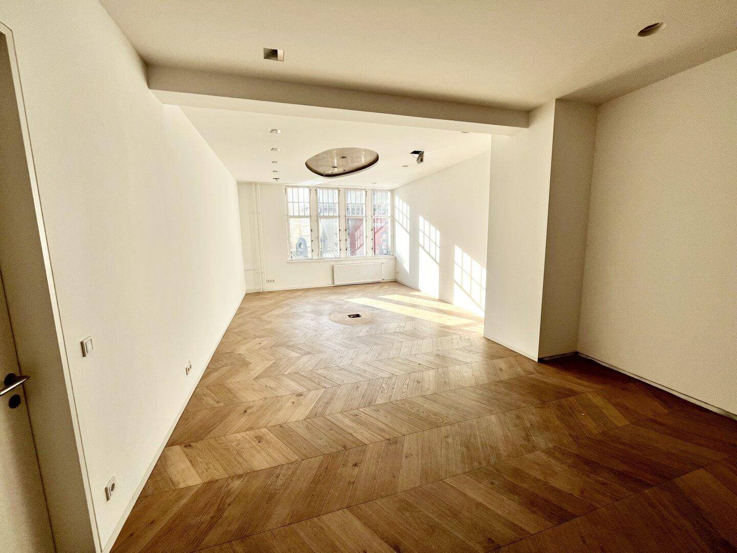Heller Raum mit Fischgrätparkett und großen Fenstern, die viel Tageslicht hereinlassen.