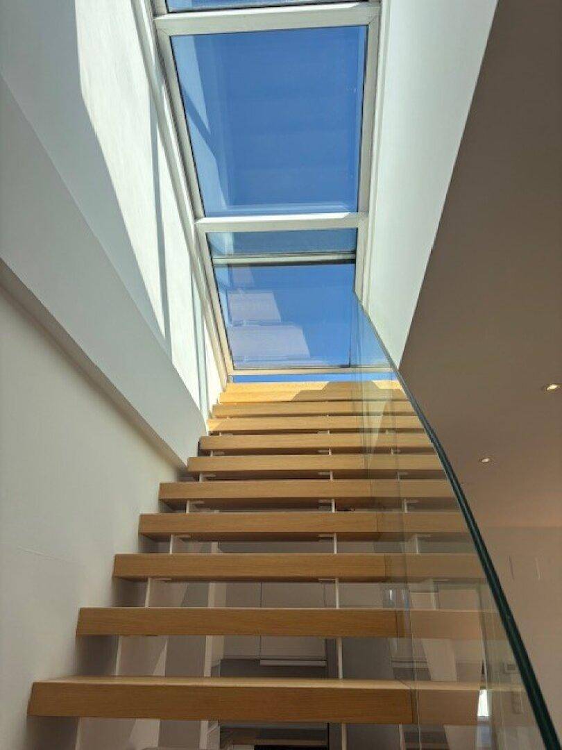 Elegante Holztreppe mit Glasgeländer, die zu einem oberen Stockwerk unter einem großen Oberlicht führt.