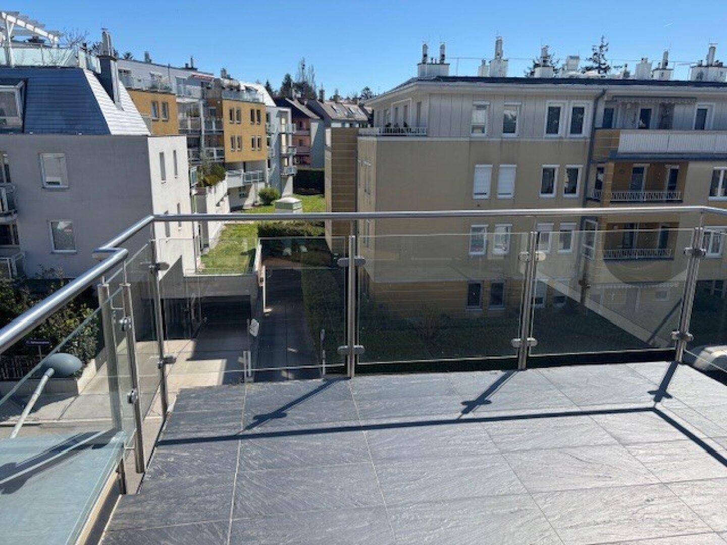 Geräumige Terrasse mit Fliesenboden und Glasgeländer, umgeben von Wohngebäuden und Grünflächen.