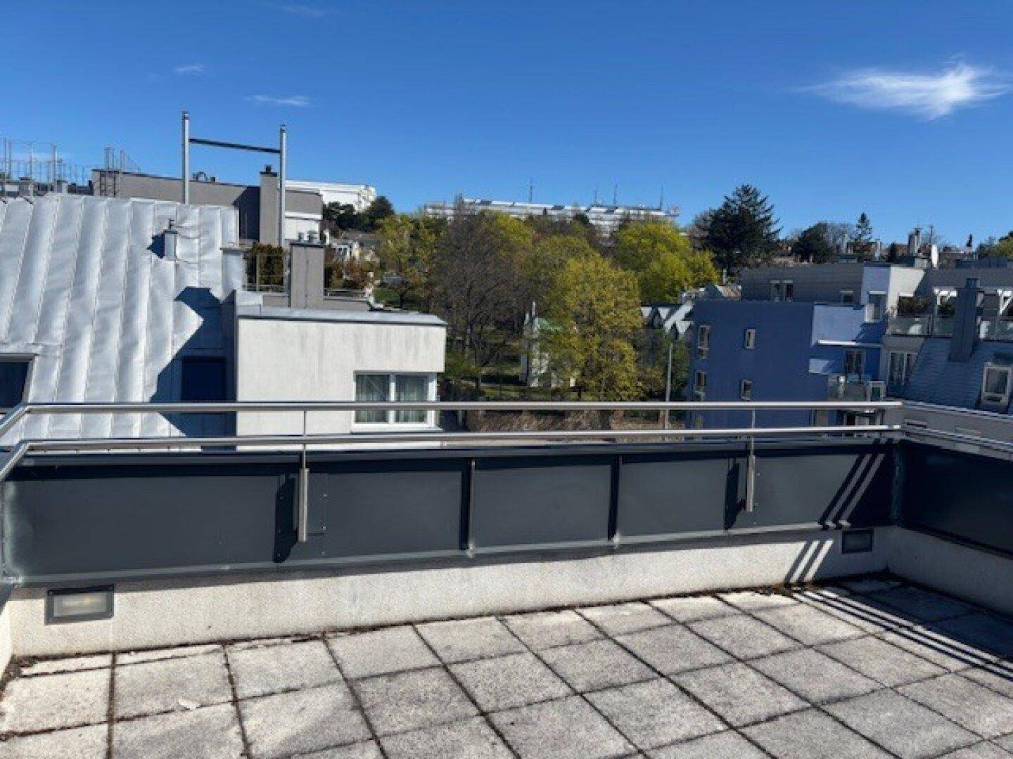 Geräumige Dachterrasse mit Fliesenboden und Metallgeländer, umgeben von Gebäuden und Bäumen.