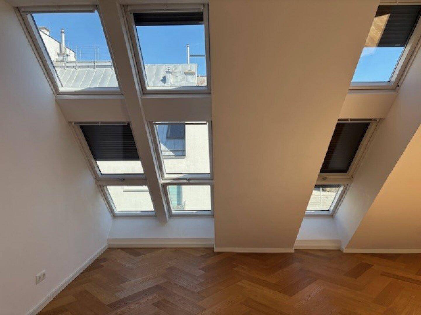 Helles Dachzimmer mit mehreren Dachfenstern und Fischgrätparkett, ideal als Schlafzimmer.